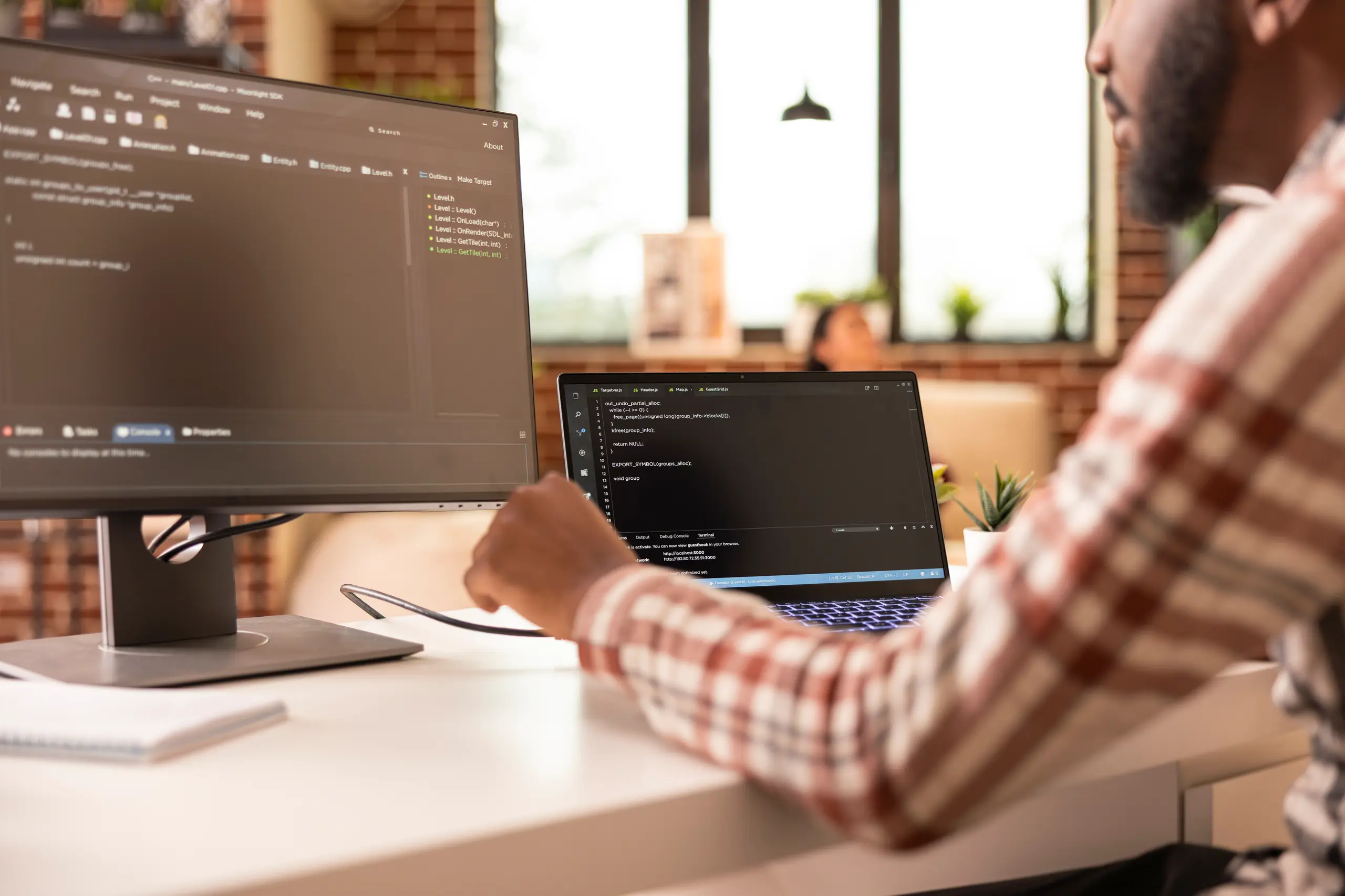 Developer at a laptop and monitor showing code in a brick-walled room